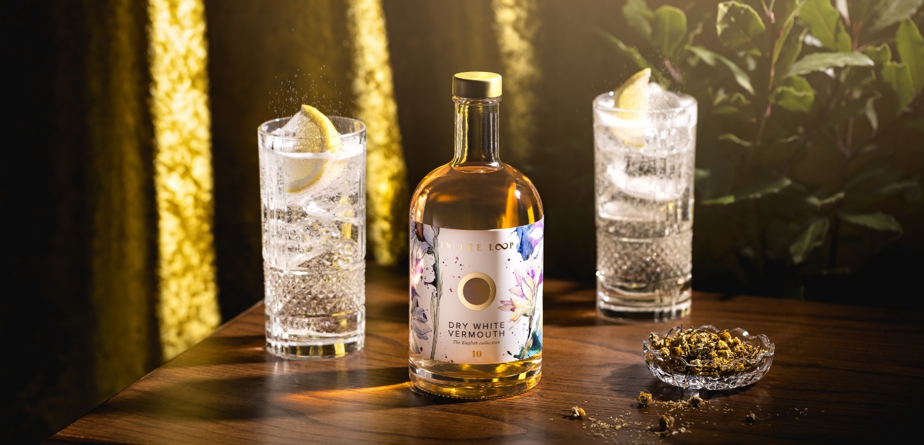 Dry white English Vermouth made and bottled in Sussex, next to Vermouth and tonic in two highball glasses with lots of ice and garnished with fresh lemon slices. Setting is gold embroidery, foliage and loose cornish chamomile